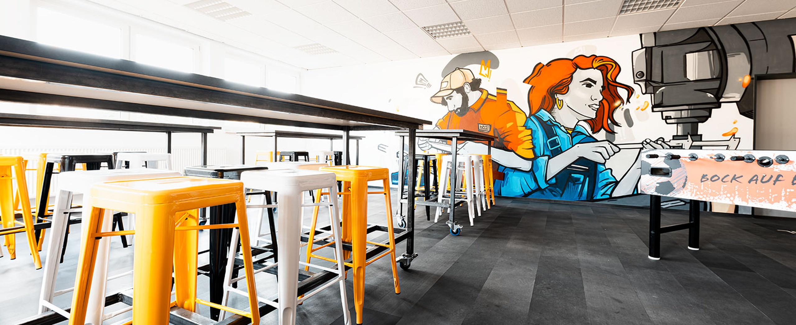 Moderner Aufenthaltsraum mit elastischem, recyceltem Bodenbelag in Schieferoptik – robust, pflegeleicht und ideal für stark frequentierte Innenbereiche wie Schulungs- oder Aufenthaltsräume. Elastischer recycelter Bodenbelag in Schieferoptik in einem modernen Aufenthaltsraum mit Hochtischen und Wandgrafik.